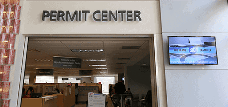 Entrance to a permit center with a sign above the doorway, staffed service desks inside, and an informational screen displayed on the wall to the right.