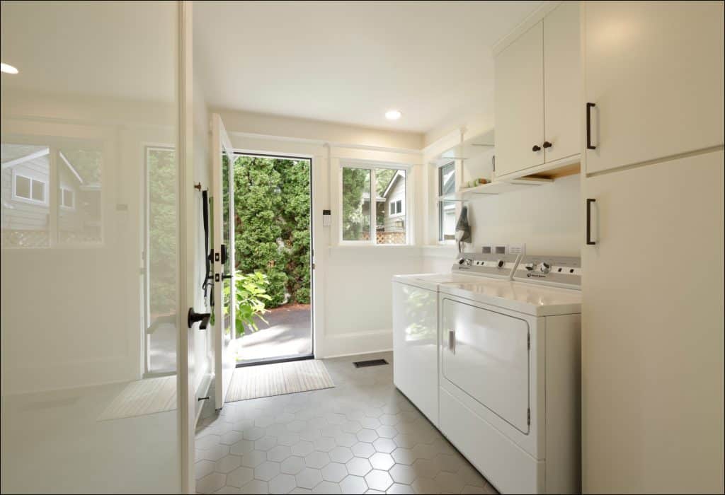 A bright laundry room, part of an award-winning residential interior remodel, features a washing machine, dryer, white cabinets, hexagonal tile floor, and a glass door that opens to a garden.