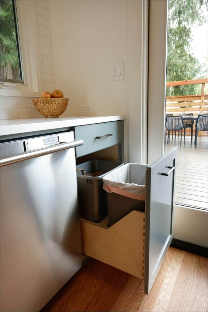 A modern kitchen, part of an award-winning residential interior remodel, features a pull-out trash and recycling drawer next to a dishwasher, with a fruit basket on the counter and a view of the deck outside.