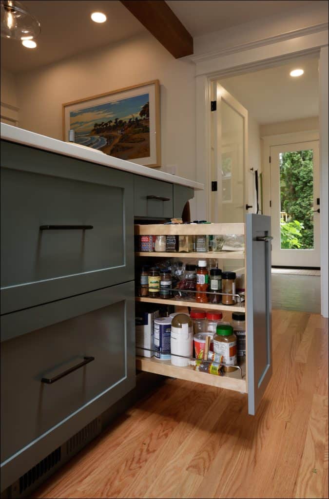 A kitchen cabinet with a pull-out shelf holds various spices and condiments in glass and plastic containers on two levels. The organized cabinet is open, showcasing the attention to detail in this award-winning remodeler of the year's residential interior.