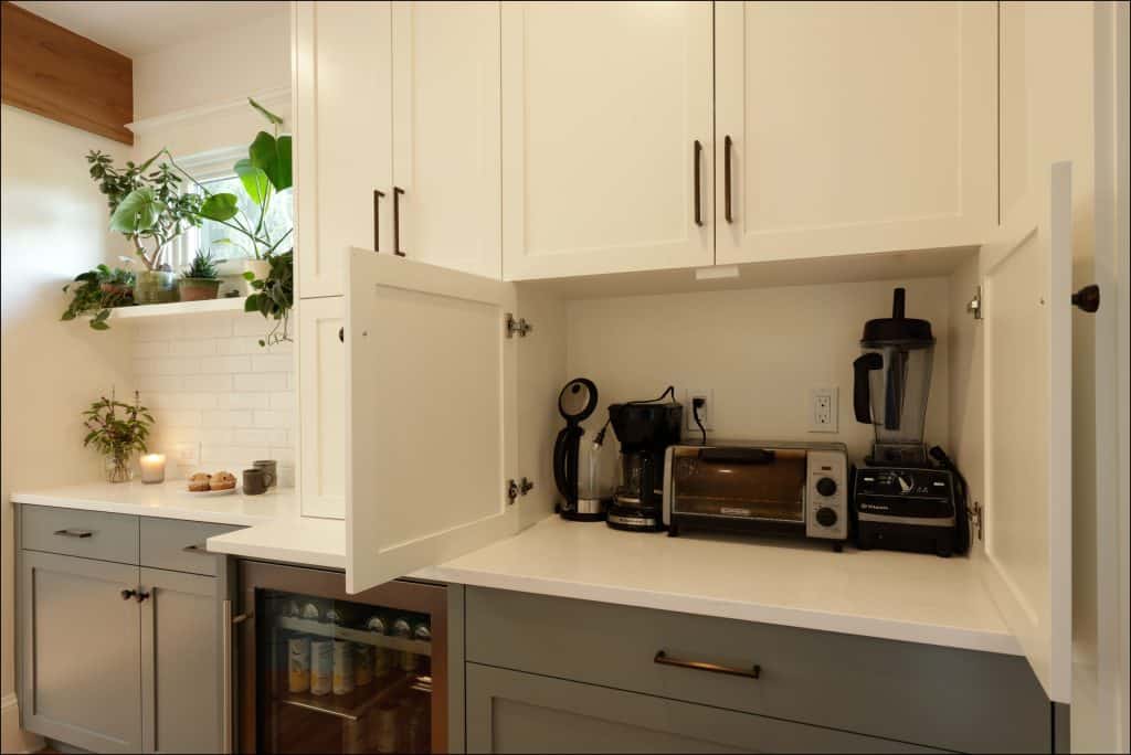 A kitchen counter with a cabinet door open reveals a coffee maker, toaster oven, blender, and toaster. Plants and a mini fridge are nearby in this award-winning residential interior.
