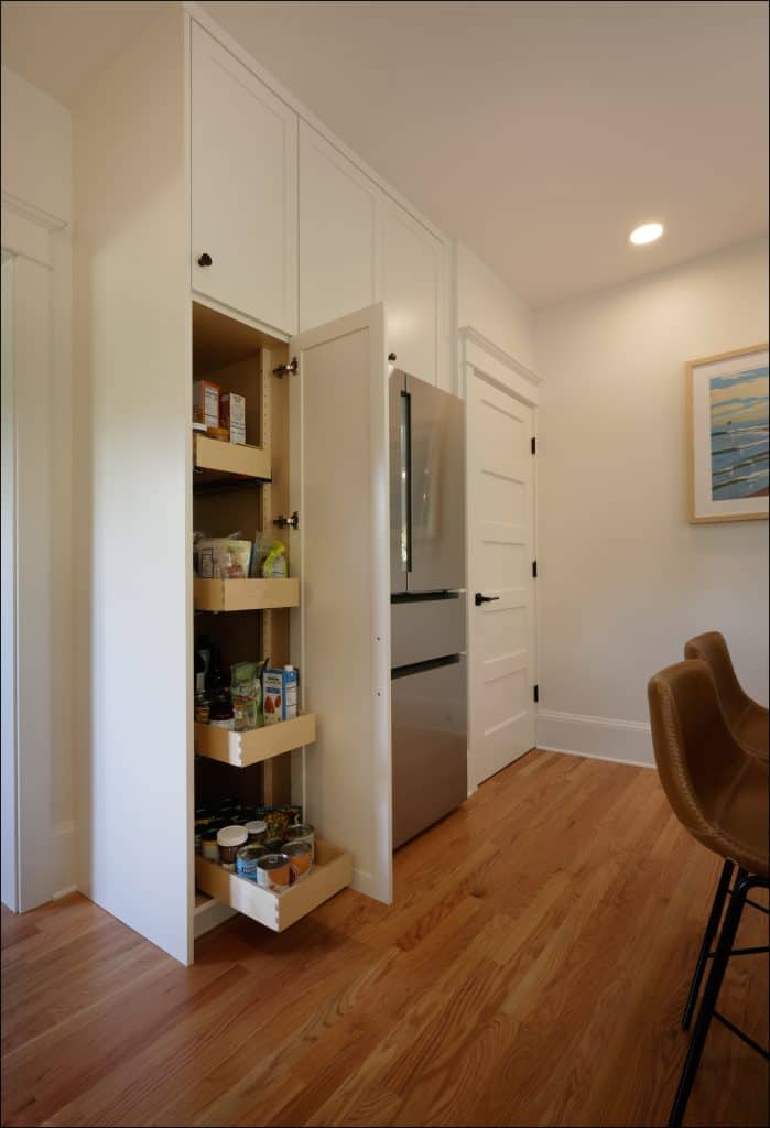 A tall white kitchen cabinet with pull-out pantry shelves is open, displaying various food items, next to a stainless steel refrigerator and brown chairs on a wood floor—part of an award-winning remodel by Remodeler of the Year.