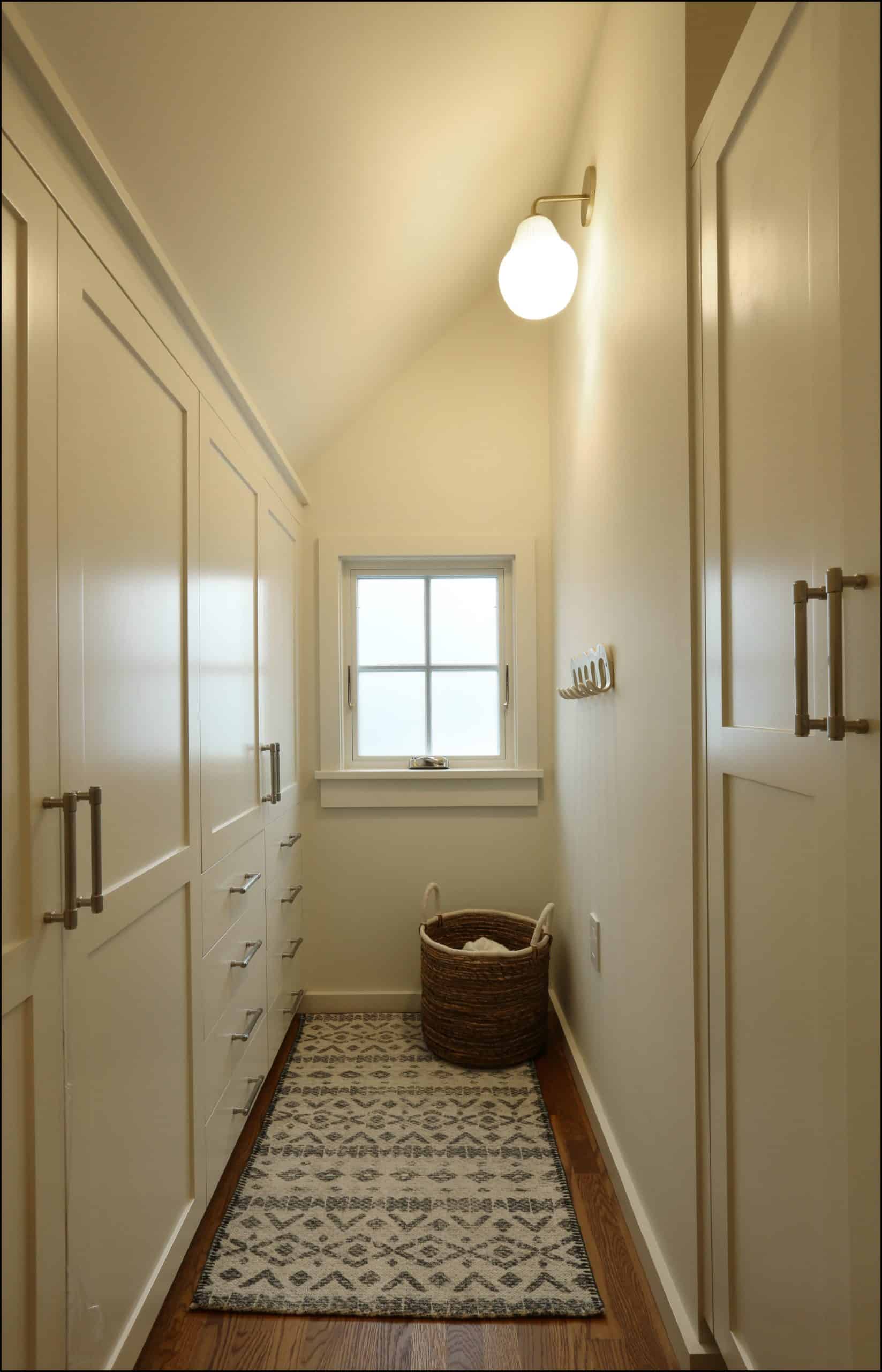 Narrow hallway with built-in cream-colored cabinets, patterned rug, wicker basket, wall hooks, and a window at the end, lit by a single wall sconce.