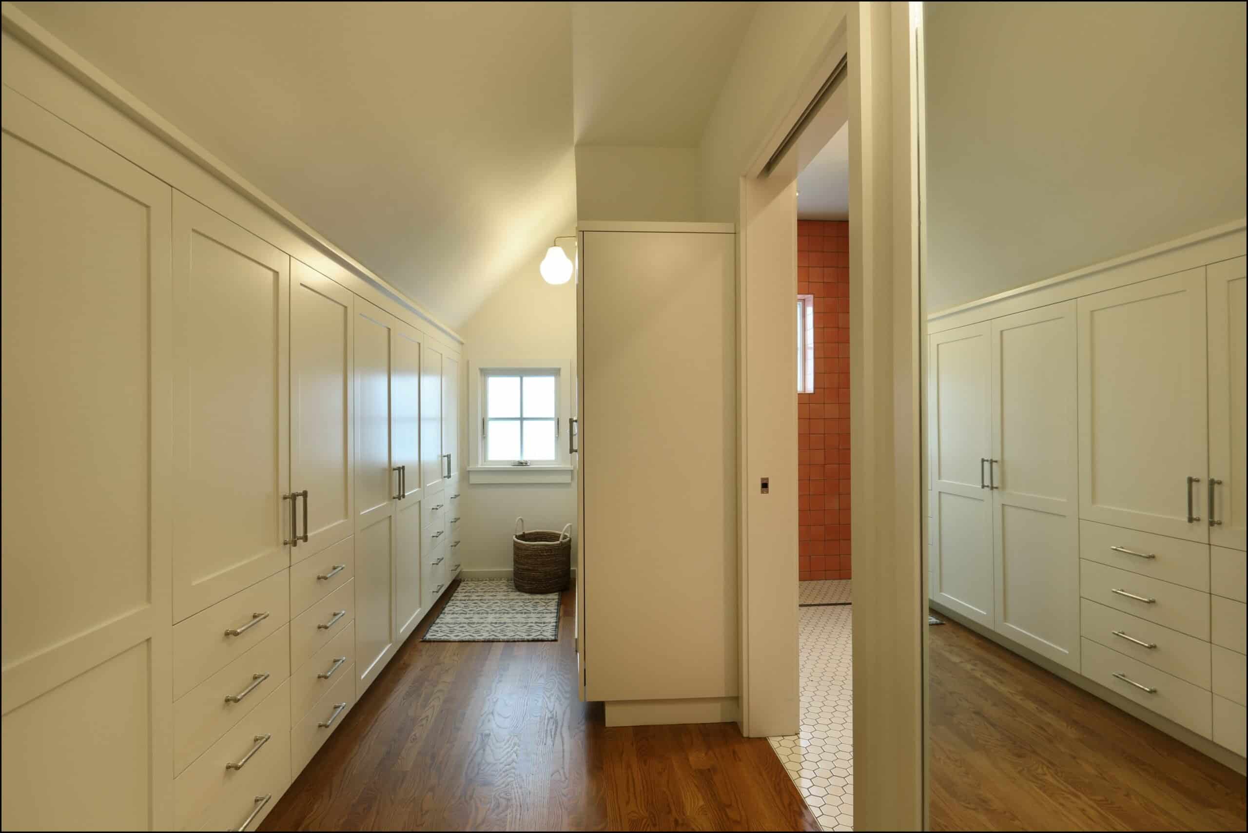 A spacious walk-in closet with built-in cabinets and drawers on both sides, wooden floor, and a view into an adjoining bathroom with tile flooring.