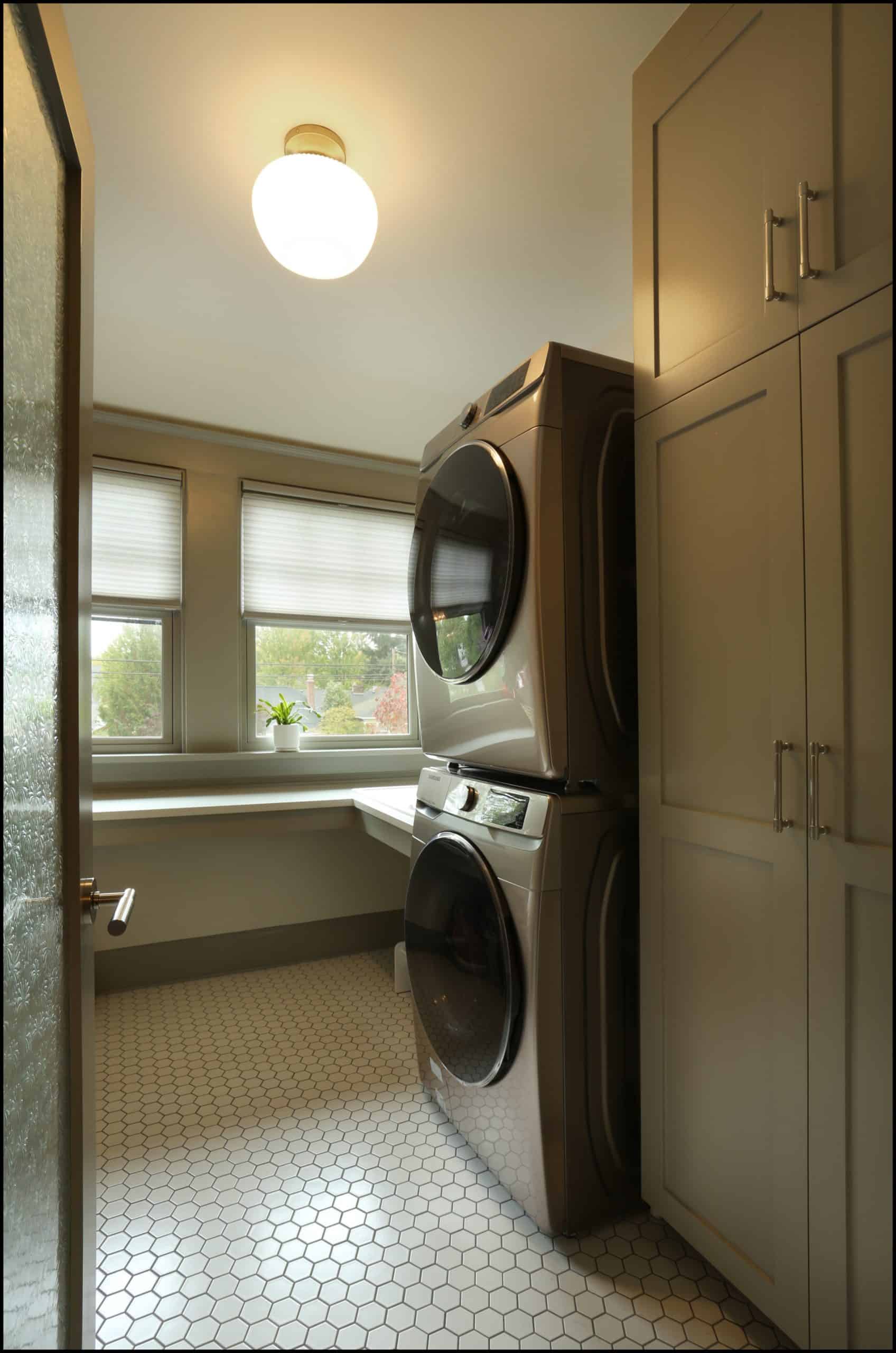 A laundry room with stacked washer and dryer units, tall cabinets, hexagonal tile flooring, and two windows with blinds.