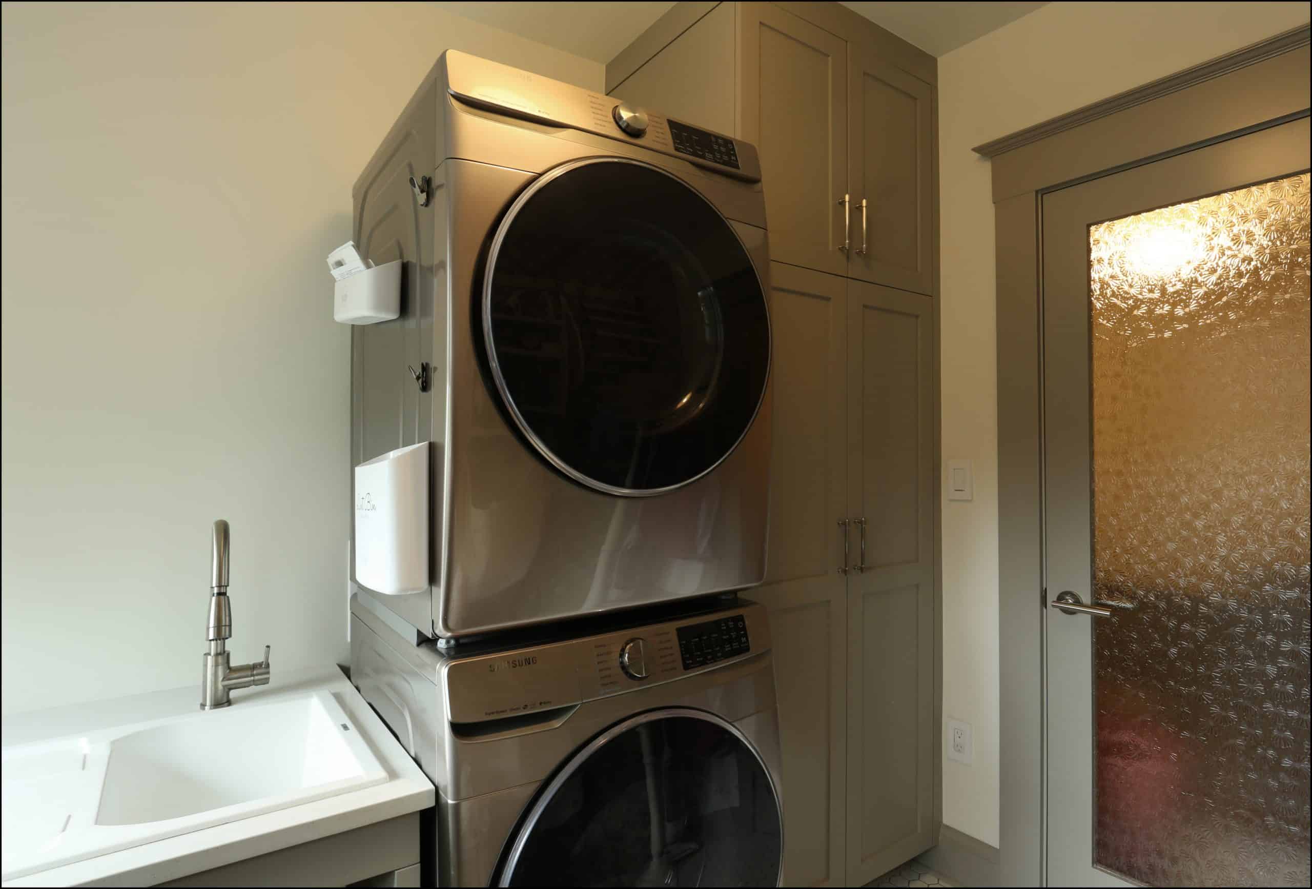 A stacked washer and dryer unit next to a utility sink and built-in cabinetry in a laundry room with a frosted glass door.
