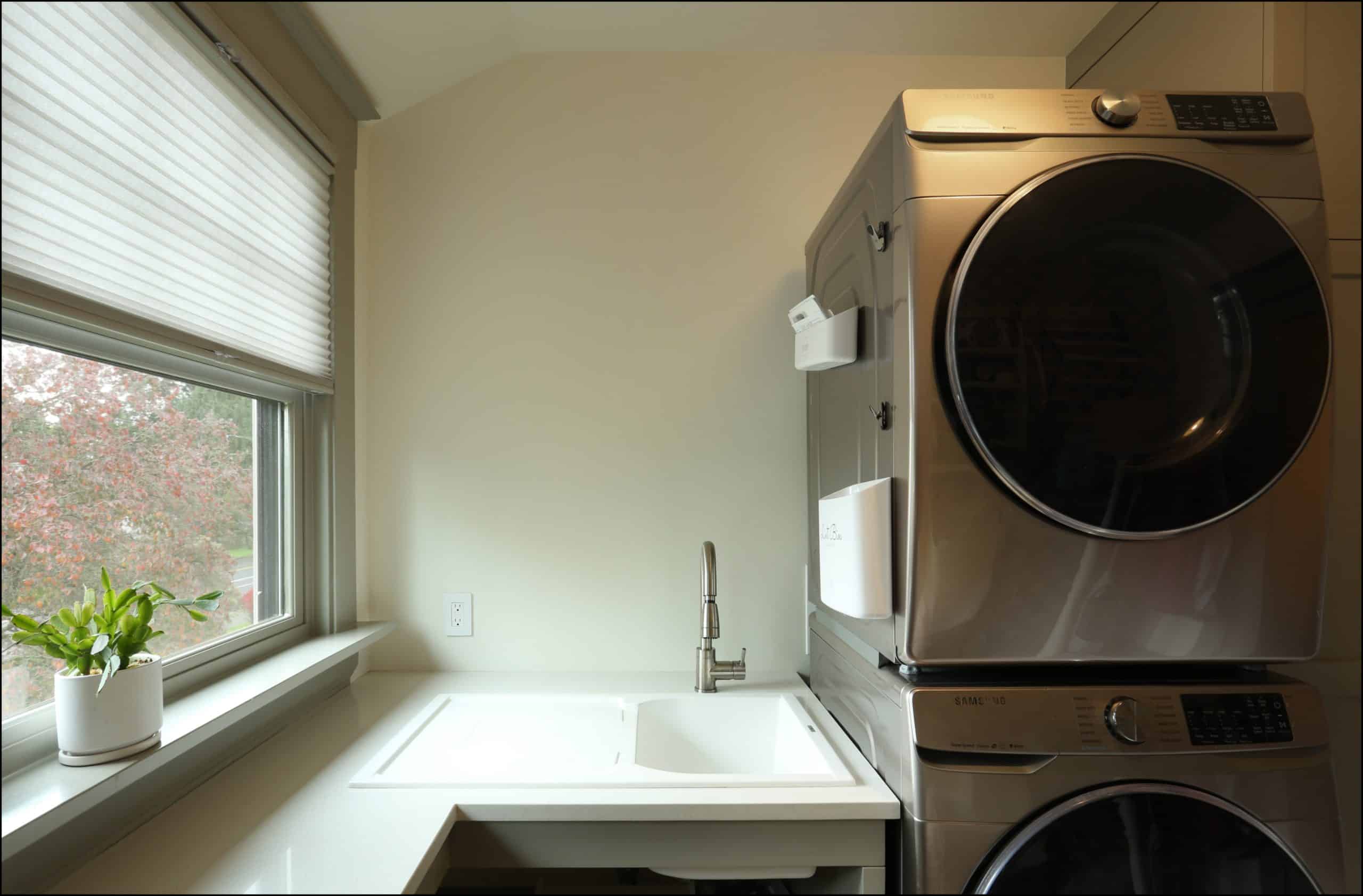 A modern laundry room with a stacked washer and dryer next to a sink, countertop, and a window with a potted plant on the sill.