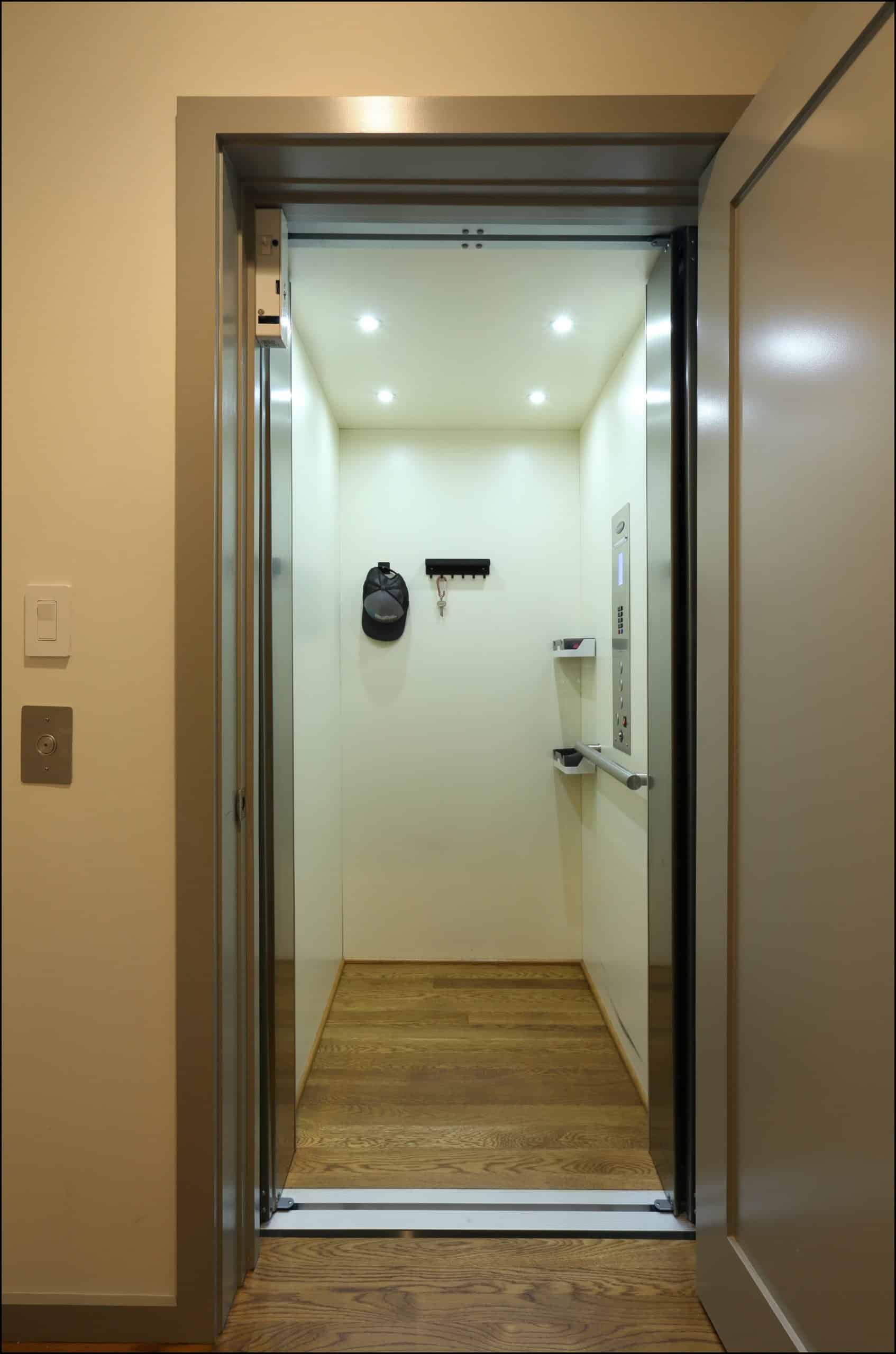 Open elevator with wood floor, cream walls, ceiling lights, control panel on the right, and a small shelf and hook on the back wall.