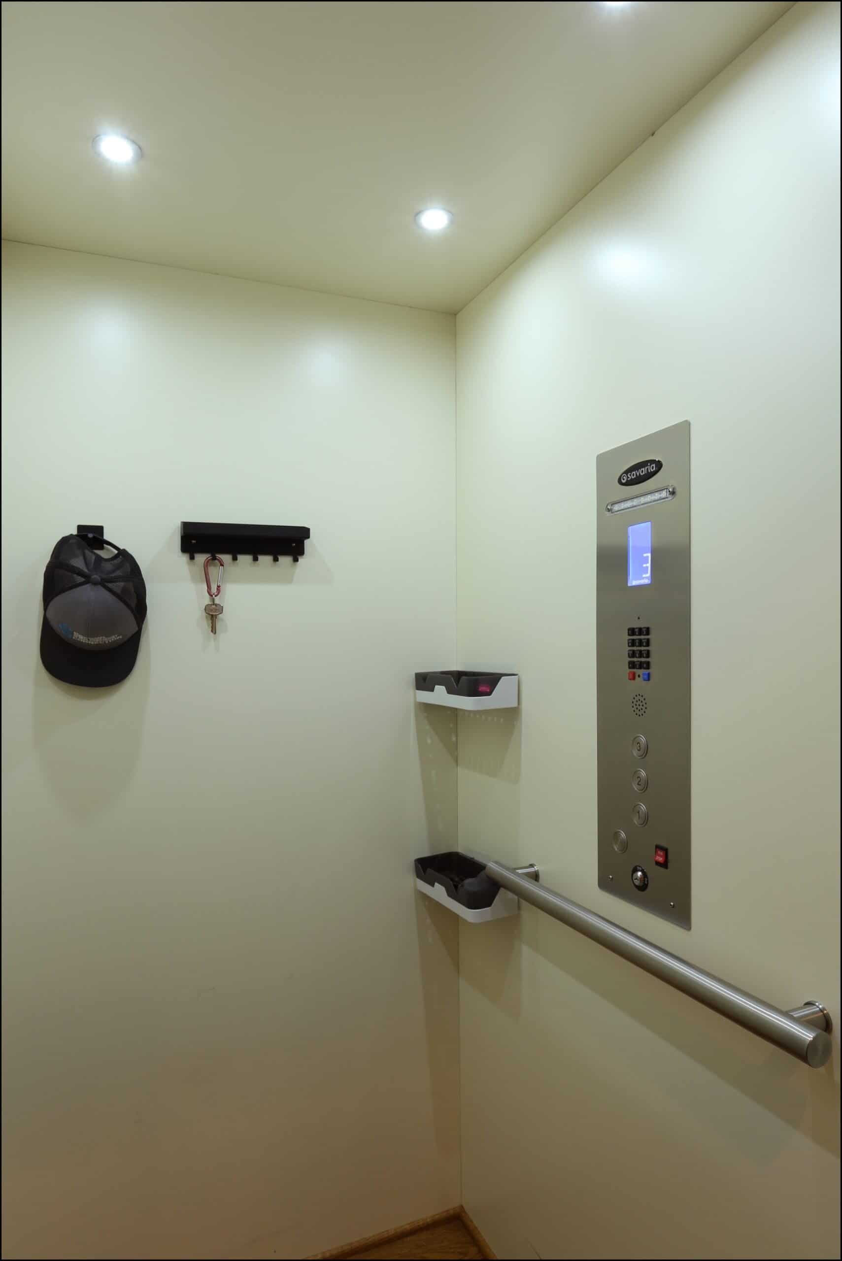 The interior of an elevator with a control panel, two small shelves, a handrail, a wall hook with keys, and a cap hanging on the wall.