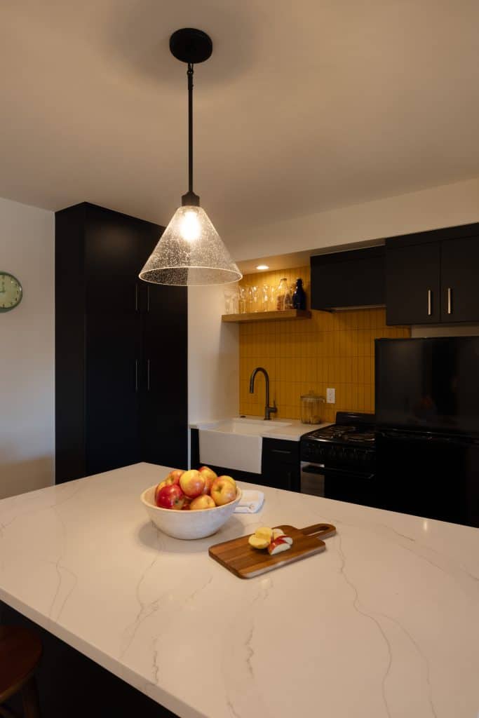 Modern kitchen with black cabinetry, yellow backsplash, and marble countertop featuring a bowl of apples and a cutting board, showcasing ReCraft Home Remodeling's design style in basement renovations.