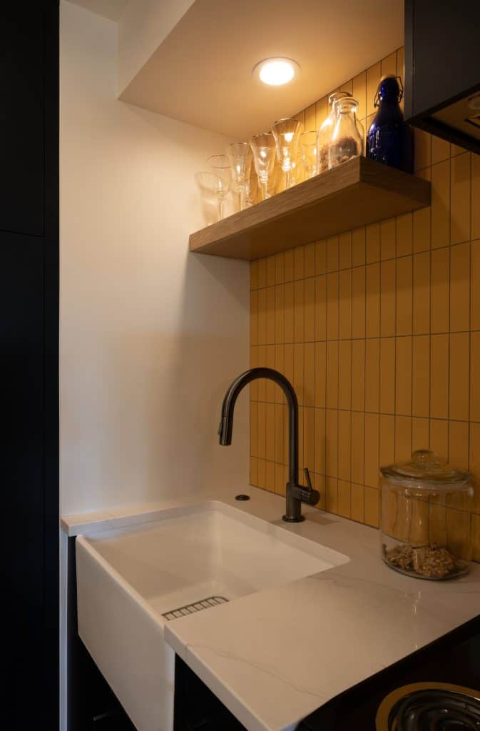 Modern basement kitchenette featuring a sleek white sink, black faucet, and a wooden shelf displaying glassware against a yellow tiled wall, showcasing ReCraft Home Remodeling's design aesthetic.