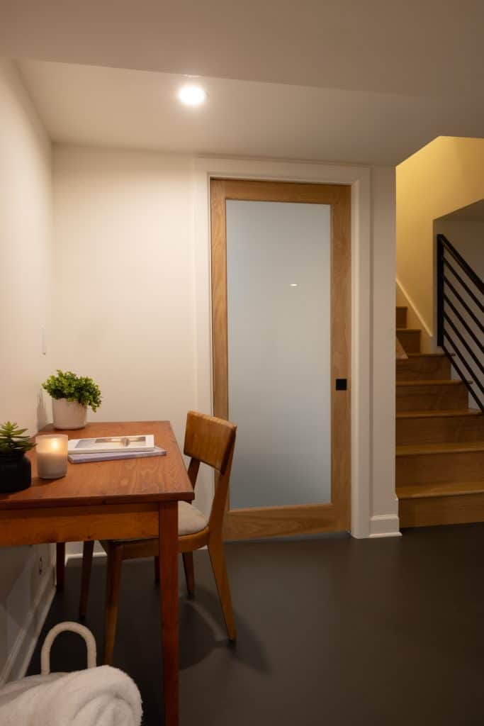 Basement remodeling project by ReCraft Home Remodeling featuring a modern workspace with a wooden desk and chair, a frosted glass door, and stairs leading to an upper level.