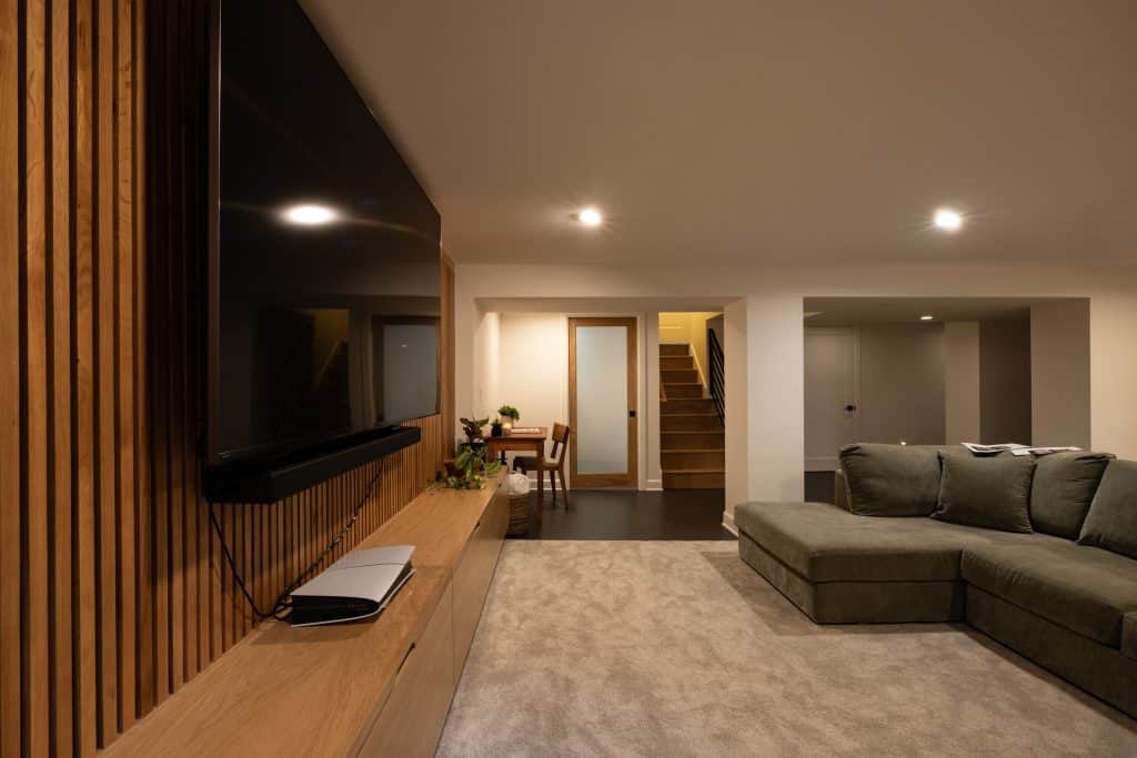 Modern basement living area featuring a green sectional sofa, wall-mounted TV, wooden accent wall, and a cozy ambiance, showcasing ReCraft Home Remodeling's design expertise in basement renovations.