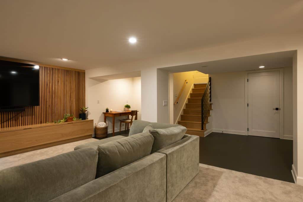 Modern basement remodel featuring a cozy seating area with a green sofa, wooden accent wall, and a TV, leading to a staircase and door, showcasing ReCraft Home Remodeling's design expertise in Portland.