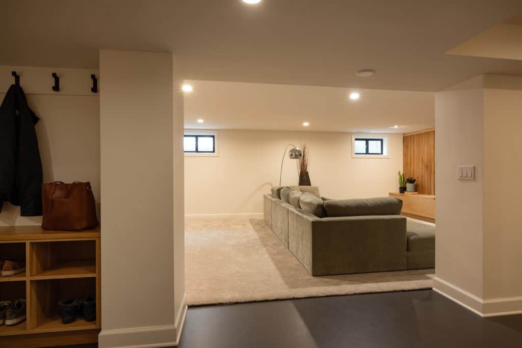Modern basement renovation by ReCraft Home Remodeling, featuring a spacious layout with a green sectional sofa, natural light from windows, and stylish decor elements.