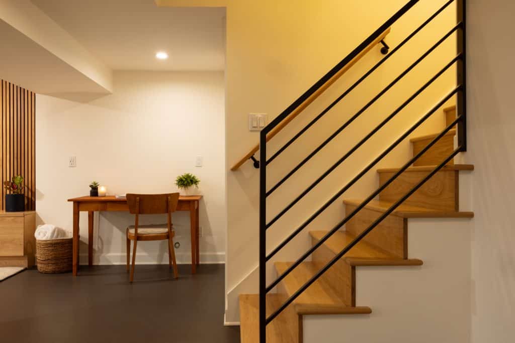 Modern basement staircase with wooden steps and black railing, leading to a well-lit area featuring a small desk and chair, showcasing ReCraft Home Remodeling's design for basement renovations in Portland.