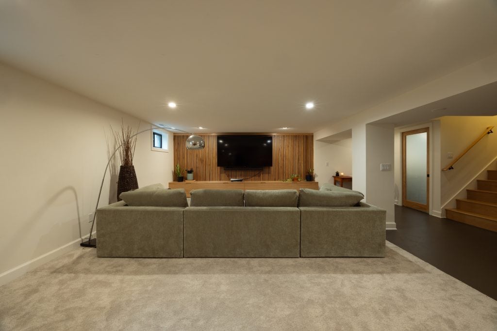 Modern basement renovation featuring a spacious seating area with a large sectional sofa, wall-mounted TV, wood accent wall, and stylish decor, showcasing ReCraft Home Remodeling's design expertise in Portland.