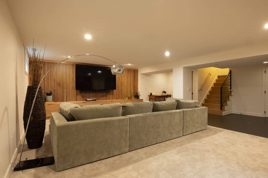 Modern basement living area featuring a spacious light gray sectional sofa, wall-mounted television, and stylish wooden paneling, showcasing ReCraft Home Remodeling's design expertise in basement renovations.