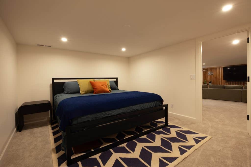 Cozy basement bedroom featuring a blue bed with colorful pillows, modern decor, and a geometric rug, showcasing ReCraft Home Remodeling's design expertise in basement renovations.