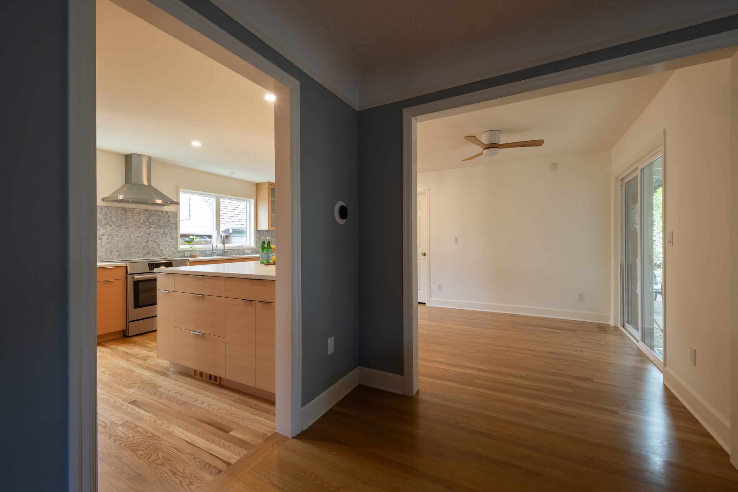 View from a hallway showing a modern kitchen with wood cabinets on the left and an empty room with wooden floors and a ceiling fan on the right.