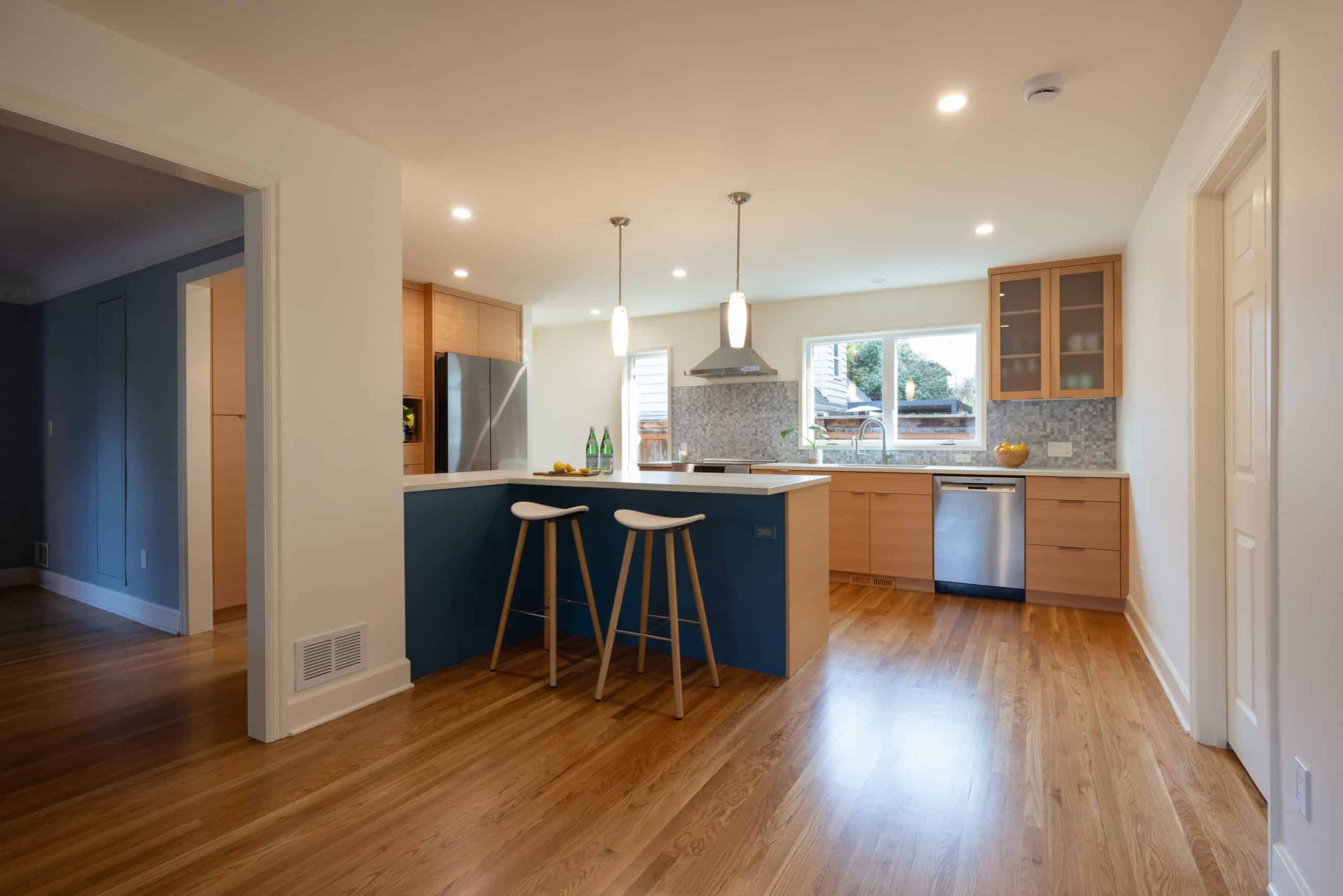 Modern kitchen with wooden floors, an island with two stools, stainless steel appliances, light wood cabinets, and pendant lighting. A window above the sink lets in natural light.