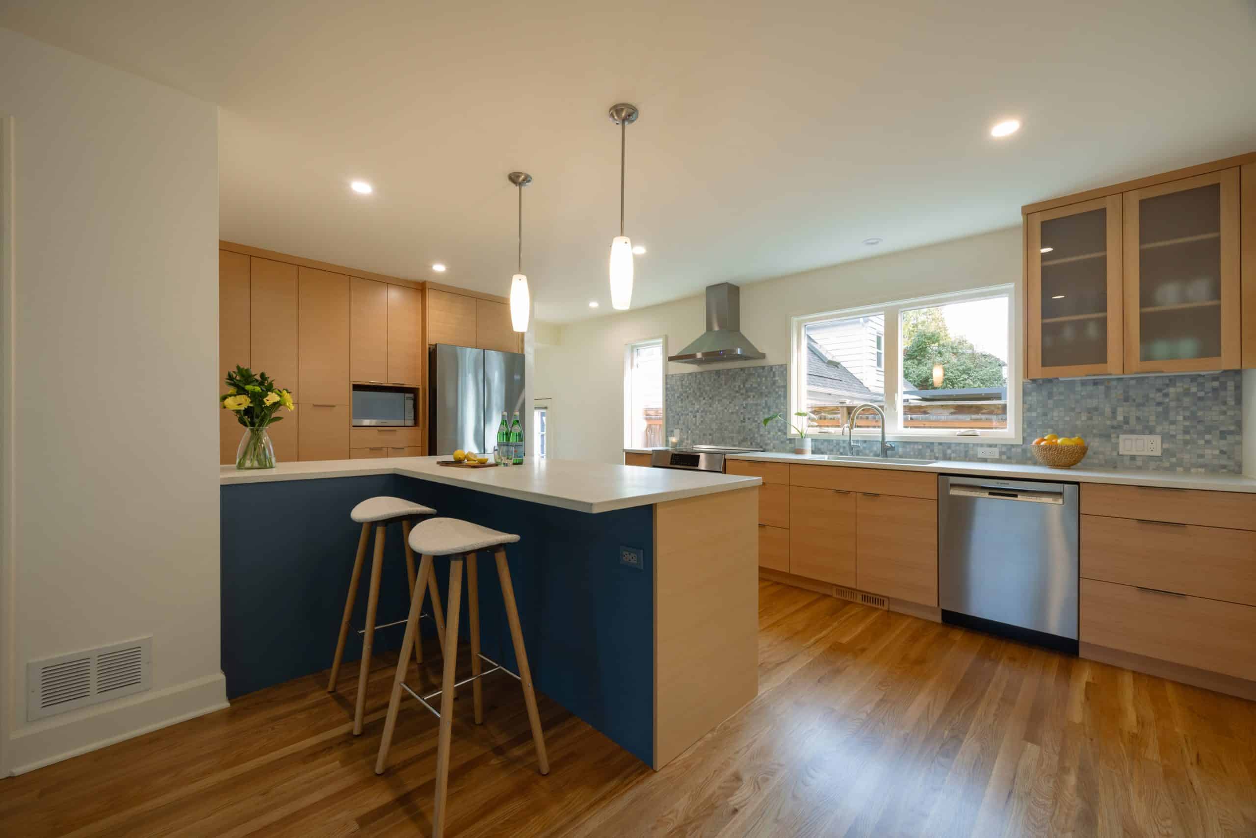 Modern kitchen with wooden cabinets, island with two stools, stainless steel appliances, pendant lights, and a window above the sink.