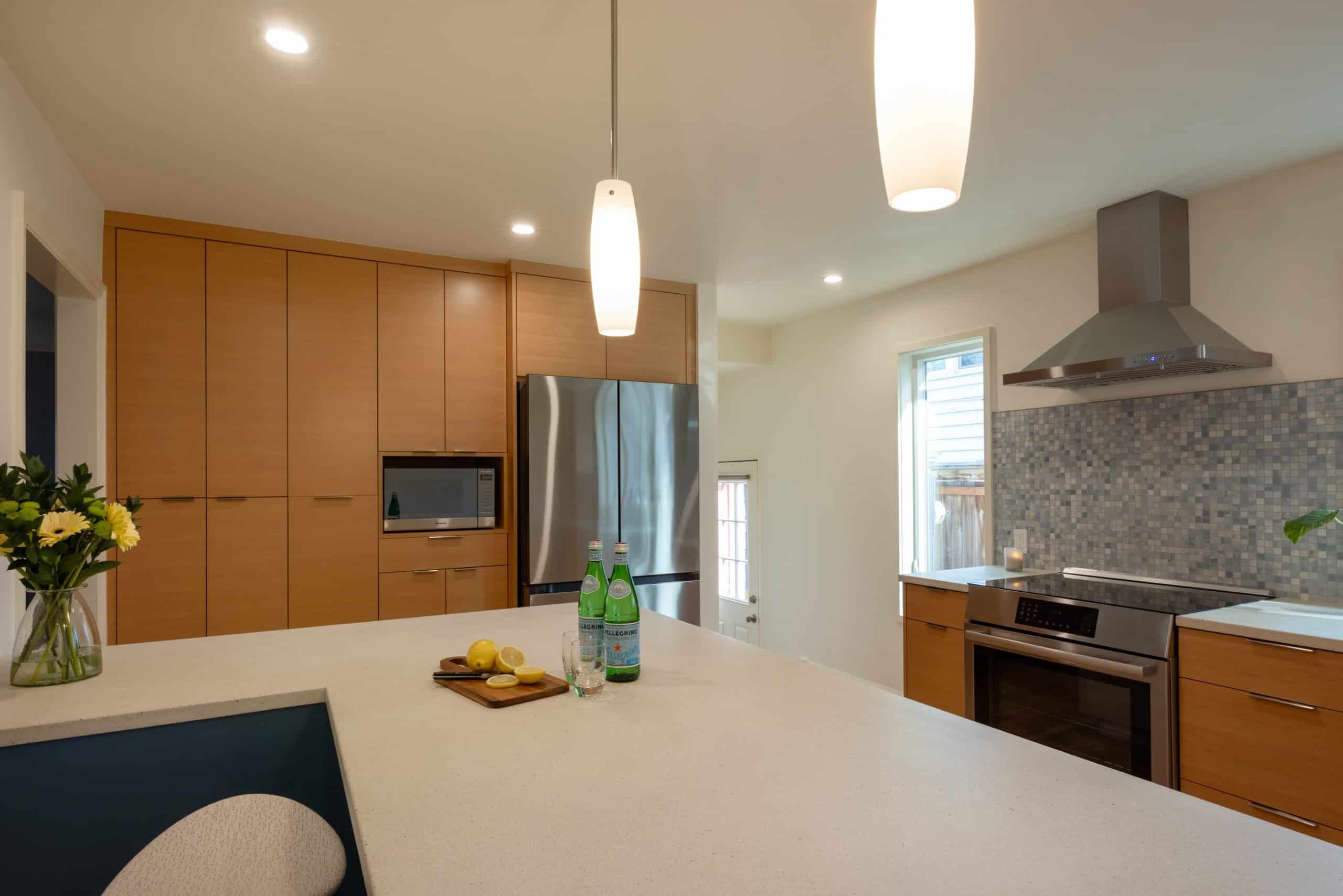 Modern kitchen with wood cabinets, stainless steel appliances, mosaic tile backsplash, and two pendant lights above a large white countertop with flowers, lemons, and bottles of sparkling water.