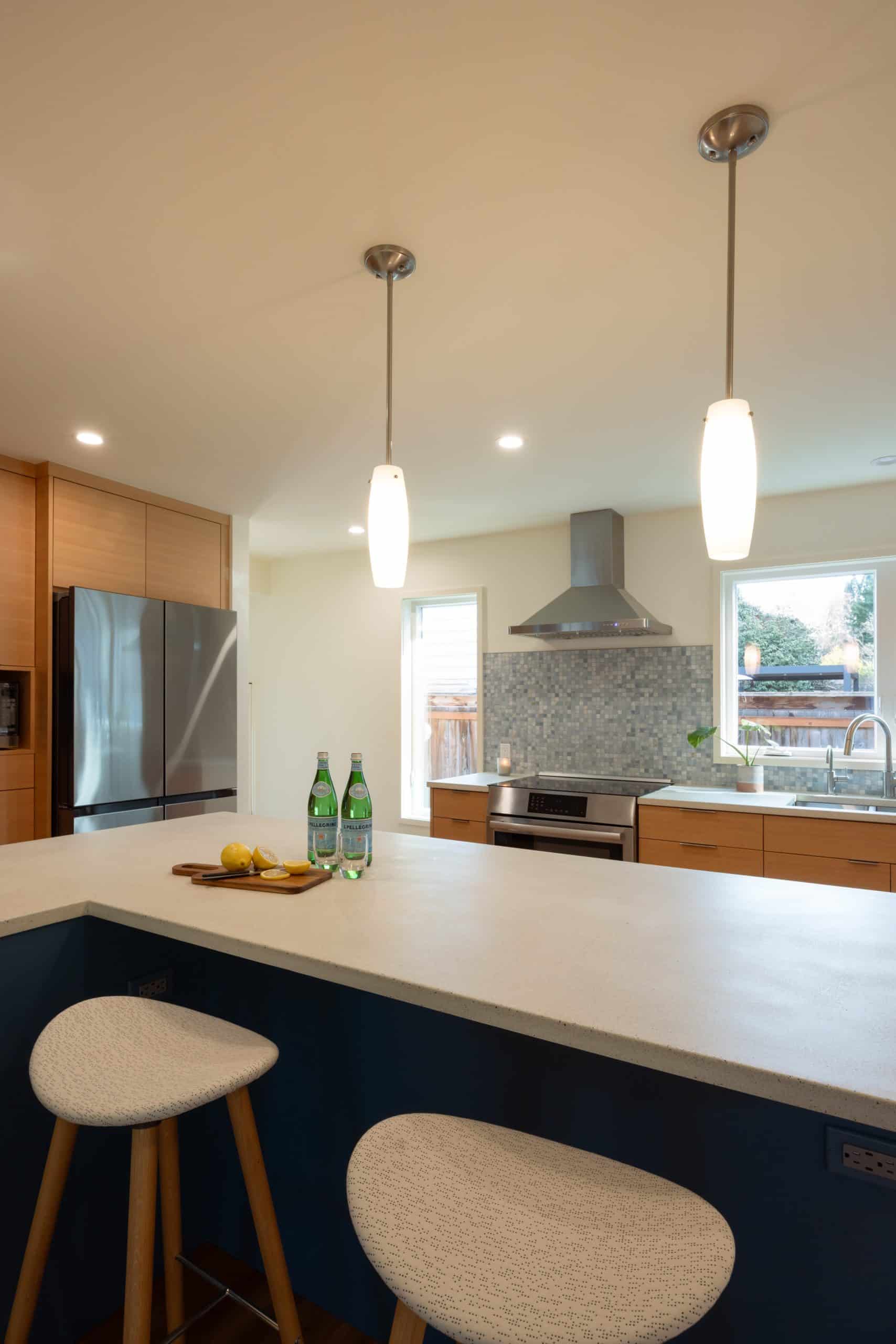 Modern kitchen with pendant lights, a tiled backsplash, stainless steel appliances, two stools at a counter, and two bottles of sparkling water with lemons on a wooden tray.