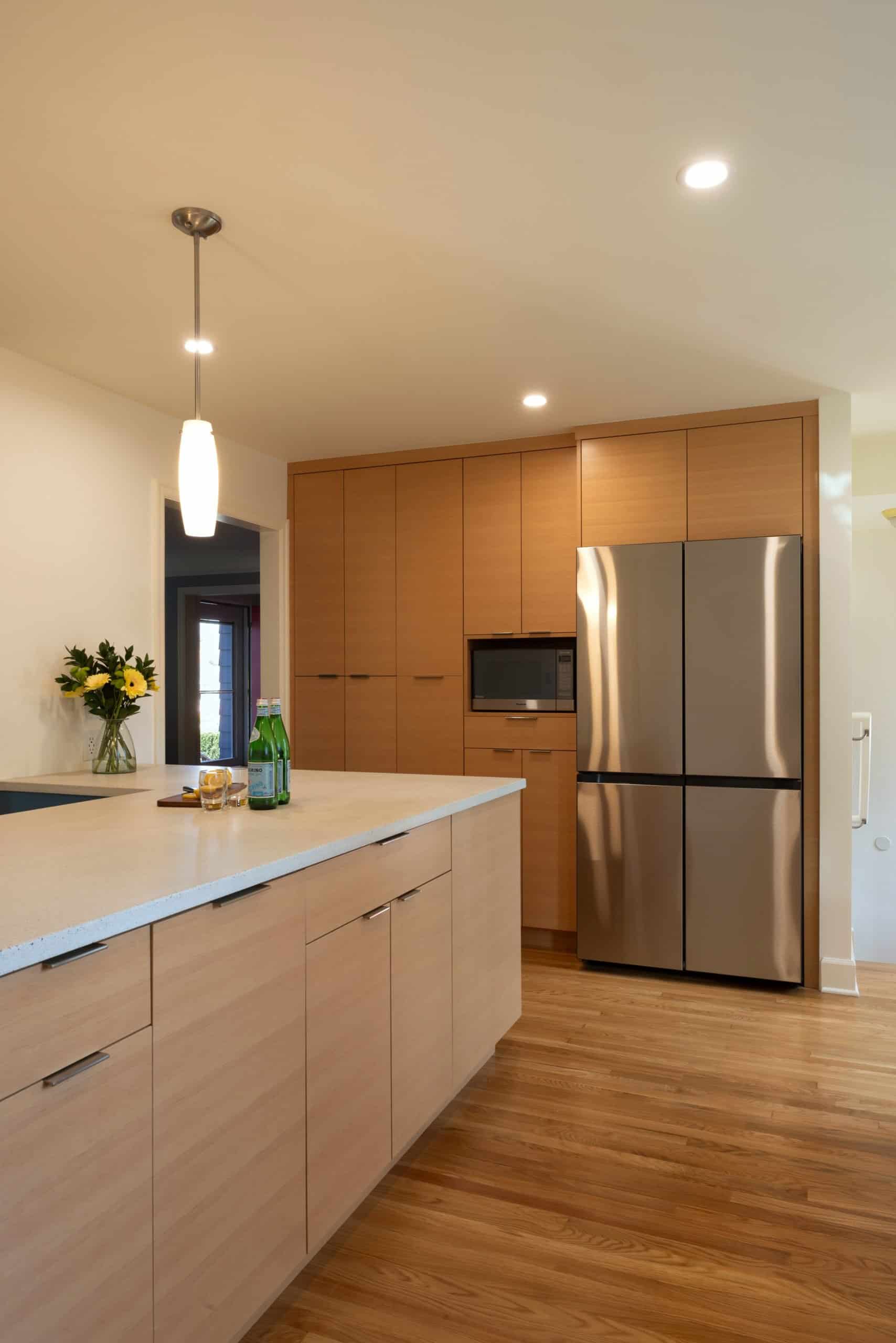Modern kitchen with light wood cabinets, stainless steel refrigerator, built-in microwave, quartz countertop island, and wood flooring. Vase with flowers and drinks on the counter.