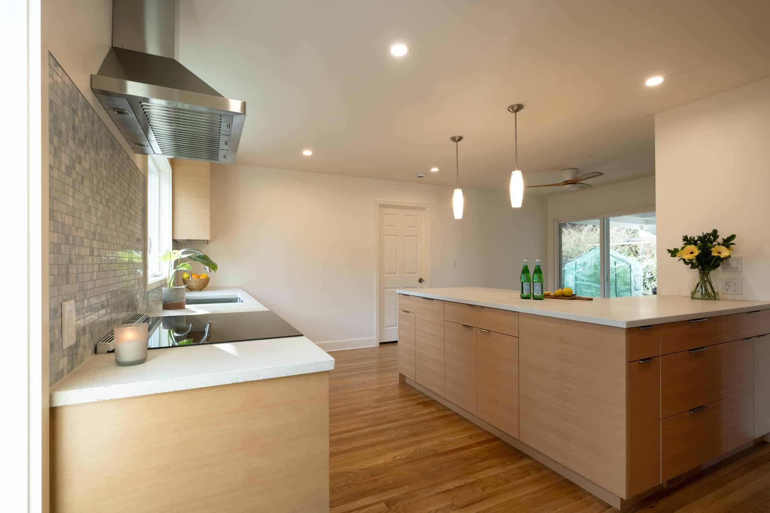 Modern kitchen with light wood cabinets, a large island, stainless steel range hood, and tile backsplash. Three bottles and a vase with flowers sit on the island.