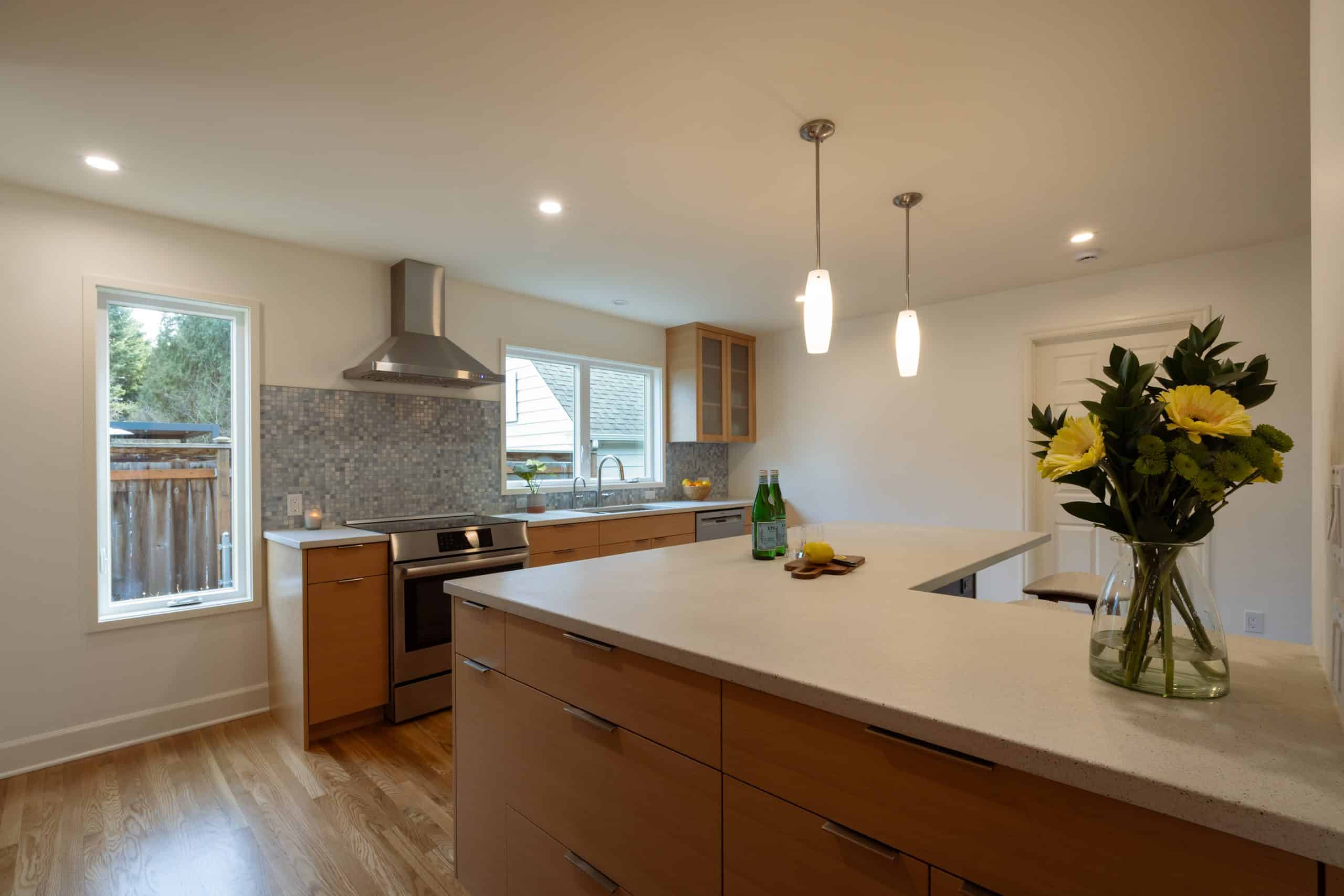 Modern kitchen with wood cabinets, stainless steel appliances, a central island with a vase of yellow flowers, and large windows providing natural light.