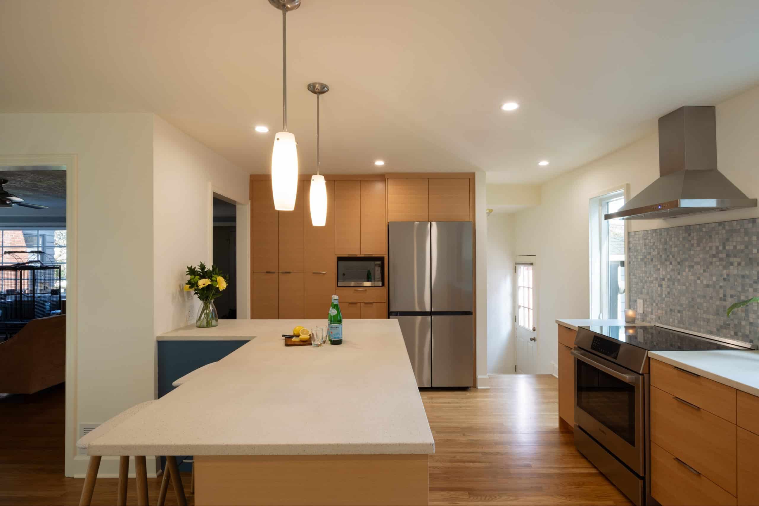 Modern kitchen with light wood cabinets, stainless steel appliances, an island with two hanging pendant lights, and a mosaic tile backsplash. A vase of flowers sits on the counter.