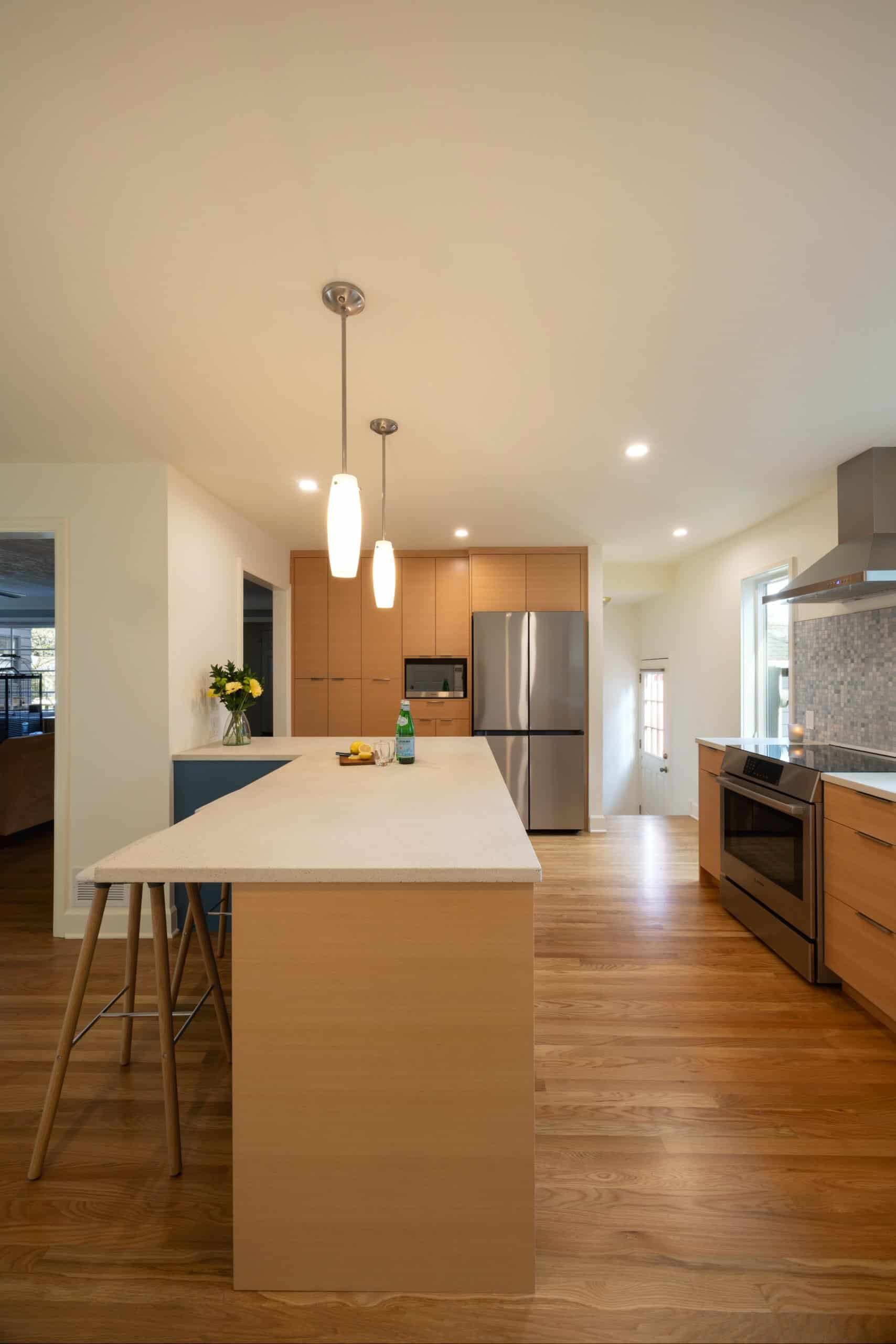 Modern kitchen with light wood cabinets, a large island with two stools, stainless steel appliances, pendant lights, and hardwood floors.