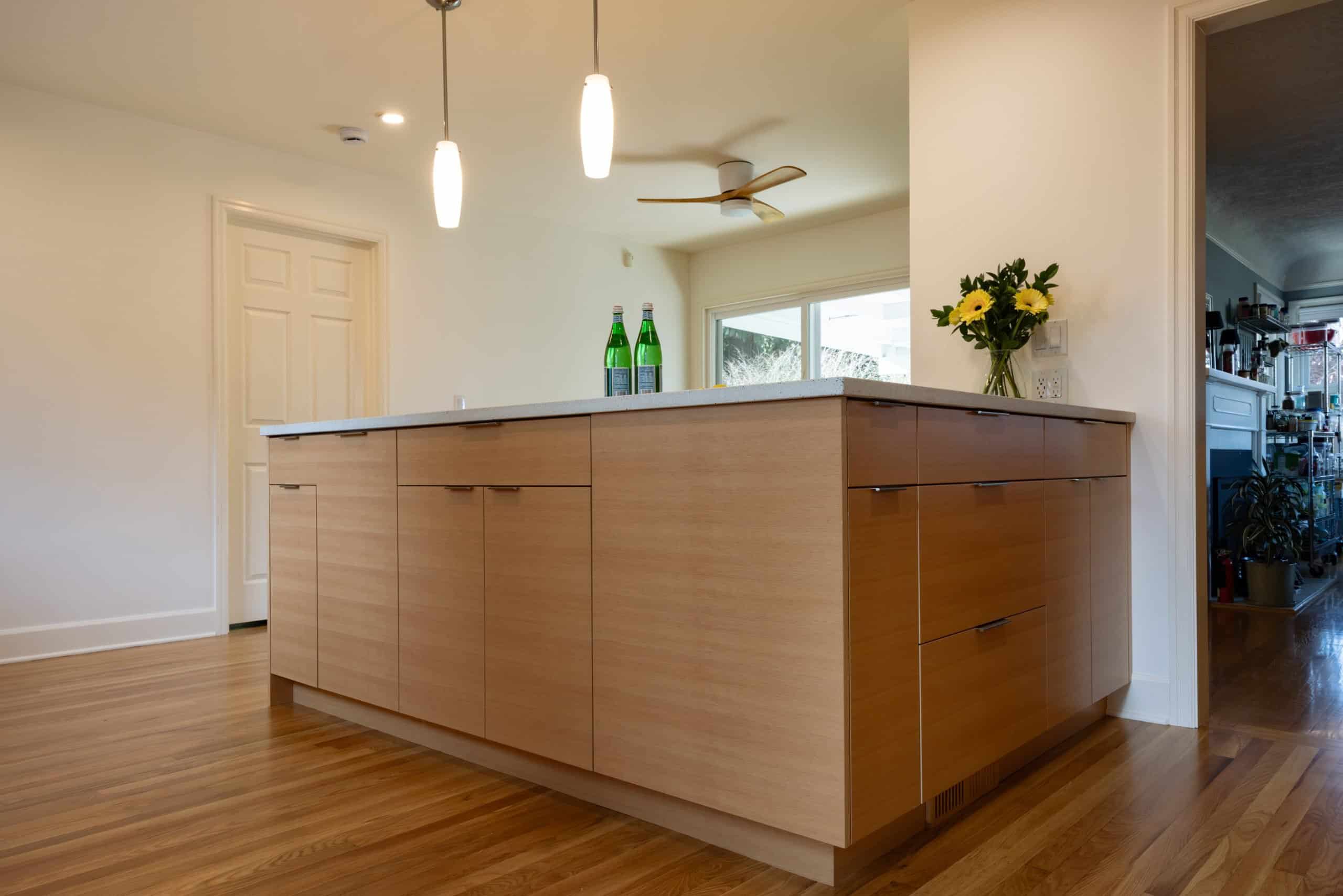 Modern kitchen with a wooden island, two pendant lights, three bottles on the counter, and a vase of yellow flowers near a doorway.