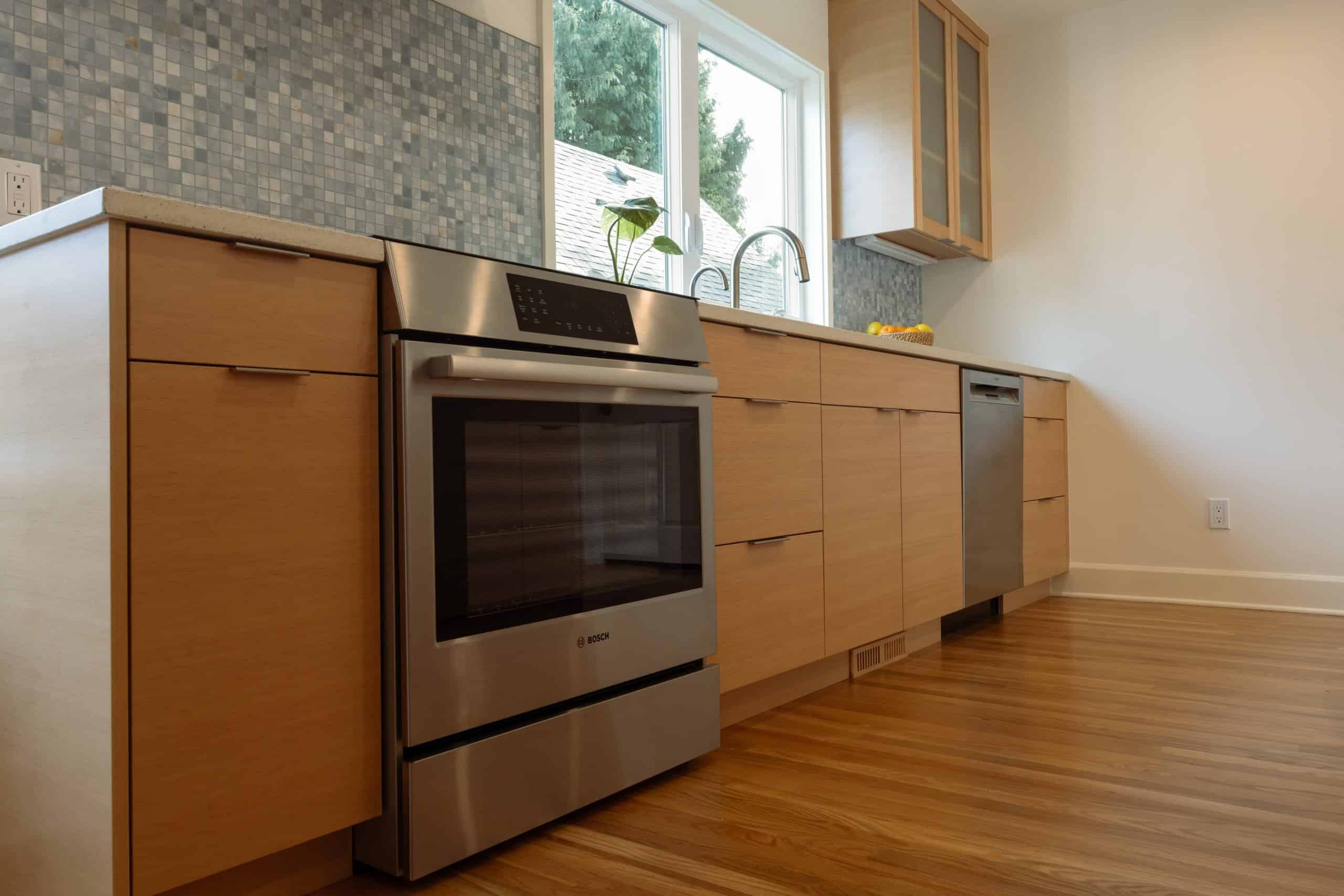 Modern kitchen with light wood cabinets, stainless steel oven and dishwasher, mosaic tile backsplash, and a window above the sink.