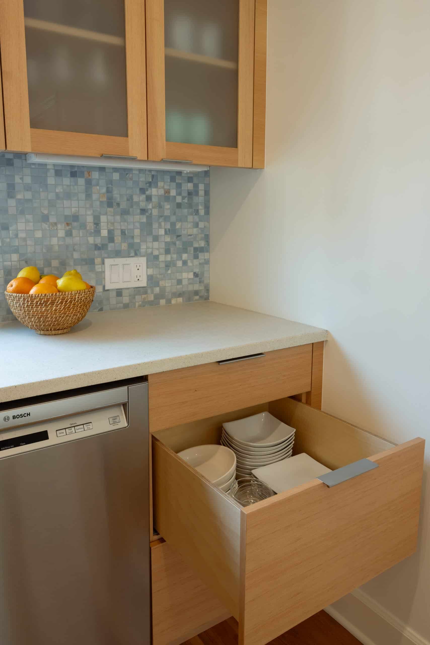 A kitchen drawer is open, showing stacked white dishes and bowls. A dishwasher is nearby, and a basket of fruit sits on the counter against a blue tile backsplash.