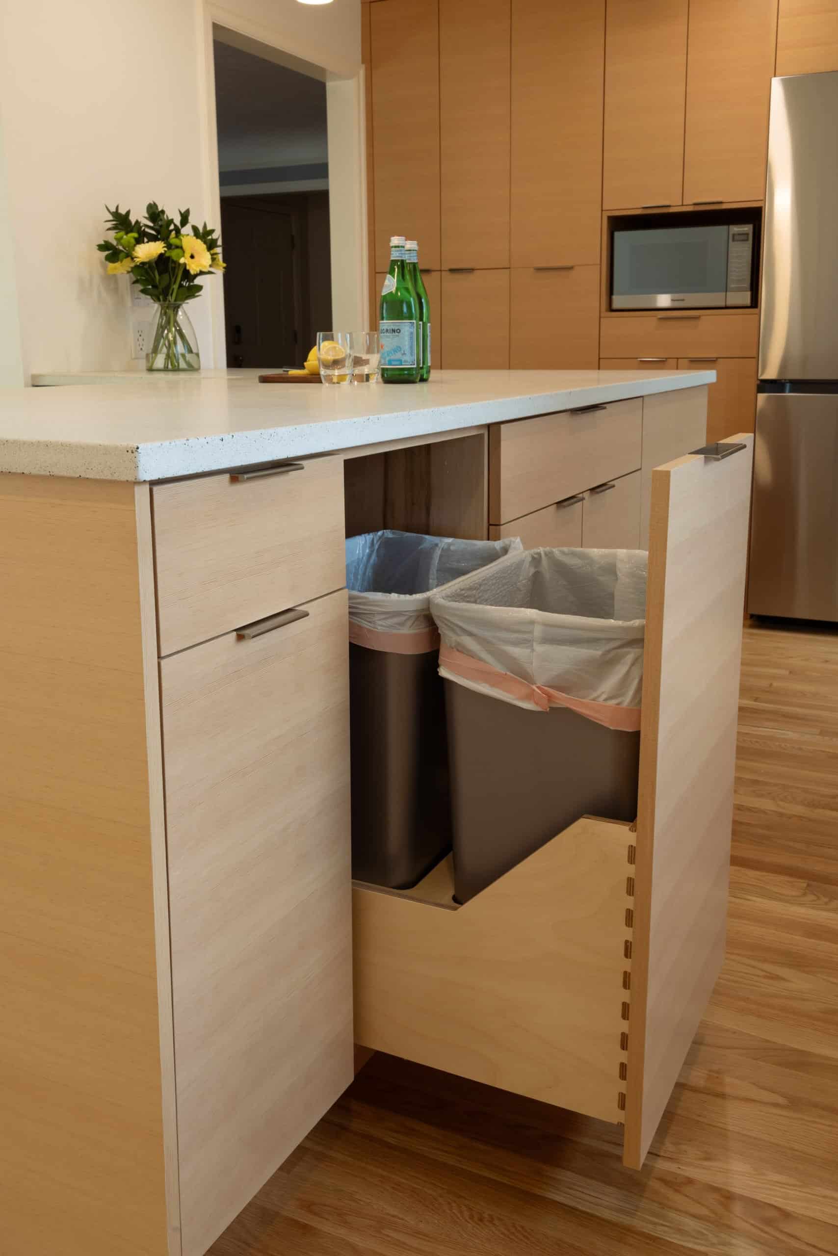 A modern kitchen with light wood cabinets features a pull-out drawer containing two trash bins; a vase with flowers and water bottles are on the counter.