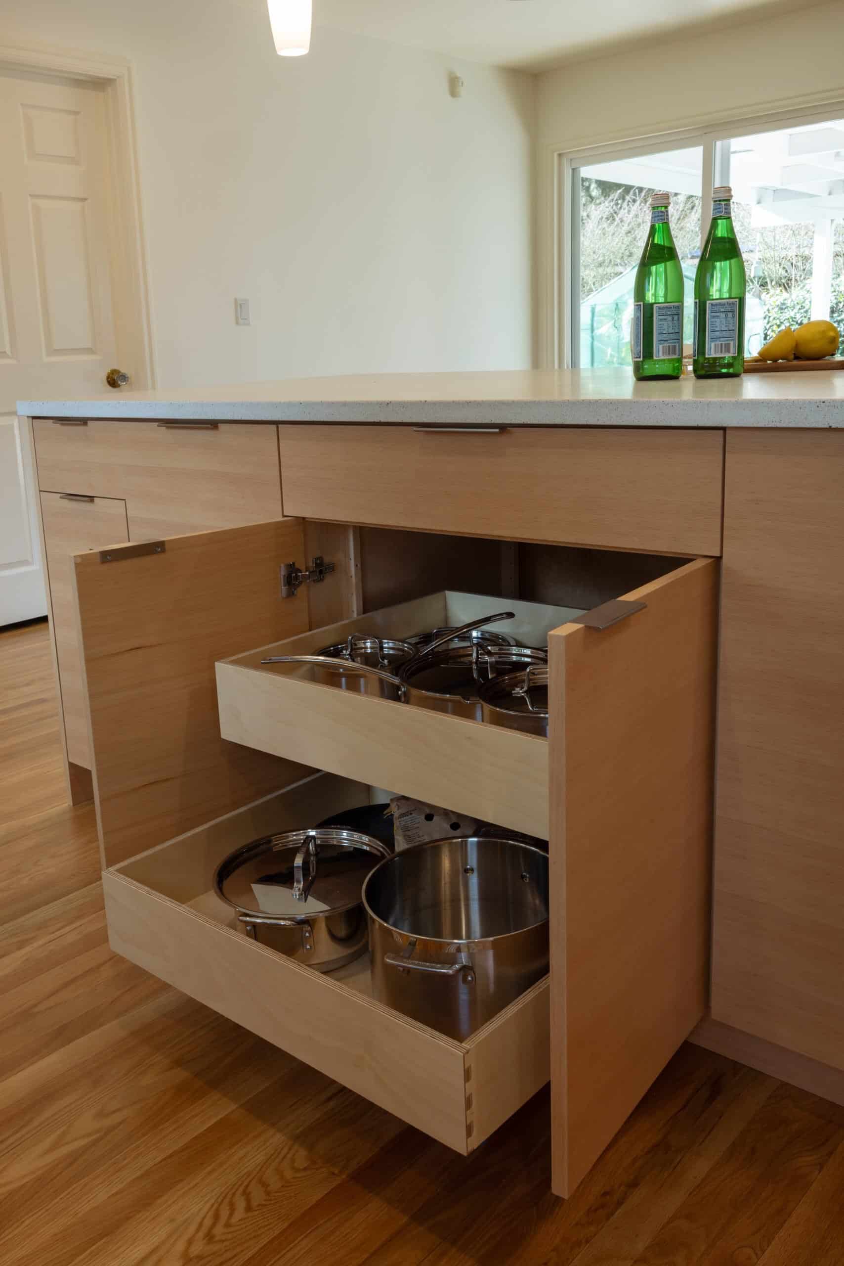 Open kitchen drawers reveal neatly stacked pots and lids on wooden shelves; two green glass bottles and a bowl of lemons sit on the countertop above.