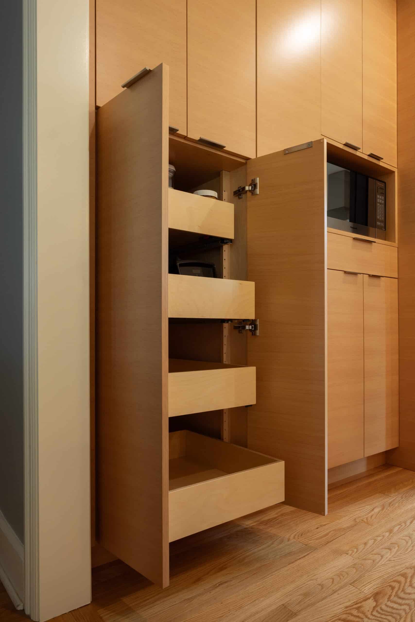 A modern kitchen with light wood cabinets, featuring an open pull-out pantry with three empty sliding shelves next to a microwave built into the cabinetry.