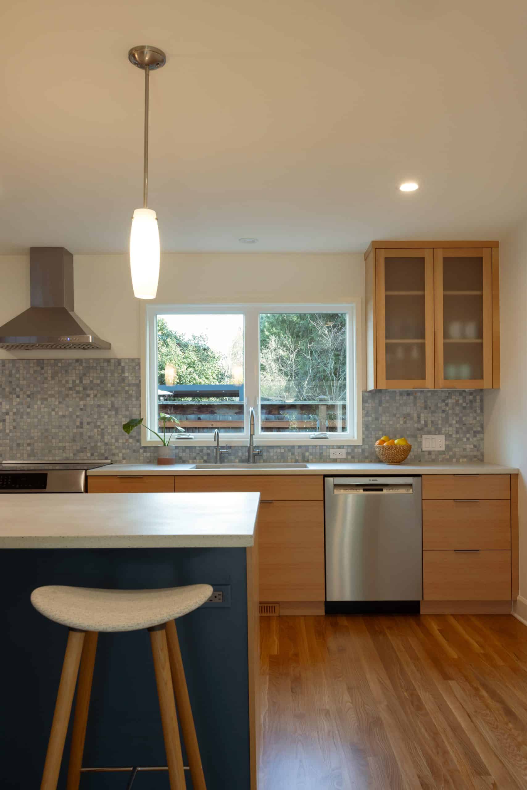 Modern kitchen with light wood cabinets, stainless steel appliances, a mosaic tile backsplash, a window over the sink, and a single barstool at a counter.