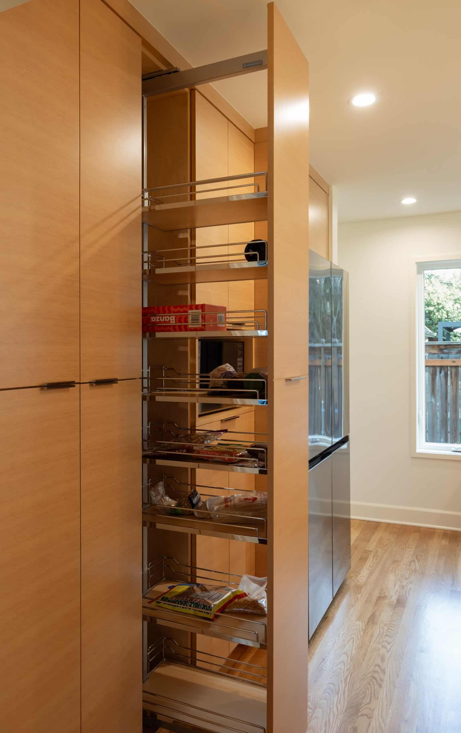 A tall wooden kitchen cabinet with a pull-out pantry drawer partially open, revealing shelves with various food items and groceries.