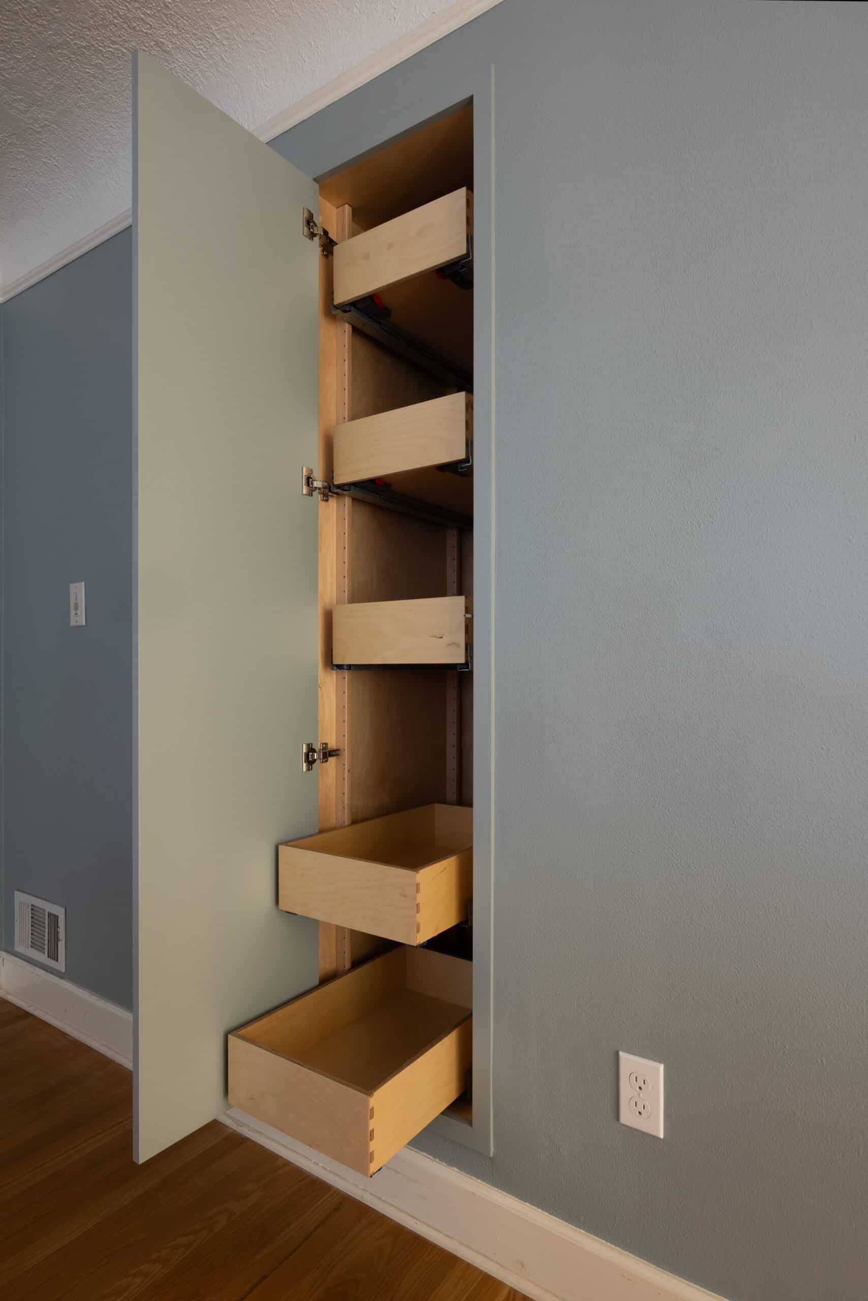 Tall cabinet with the door open, revealing four sliding wooden drawers inside, built into a light blue wall above a hardwood floor.