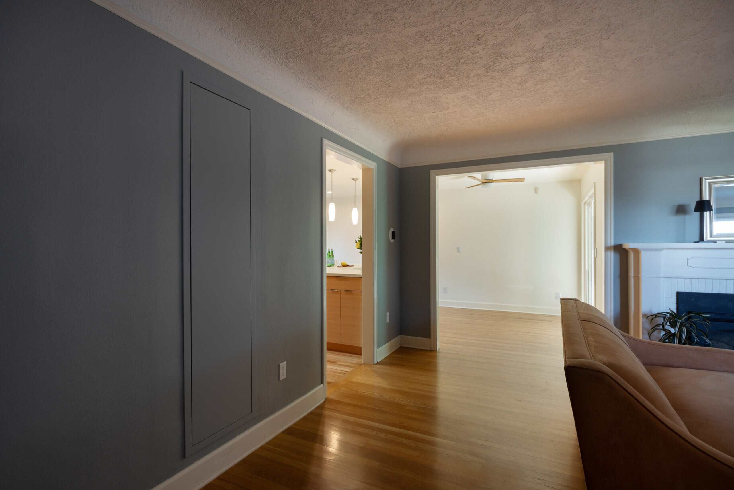 Hallway with light wood flooring leads to a kitchen on the left and an open living space with a ceiling fan and fireplace on the right. A tan sofa is partially visible.
