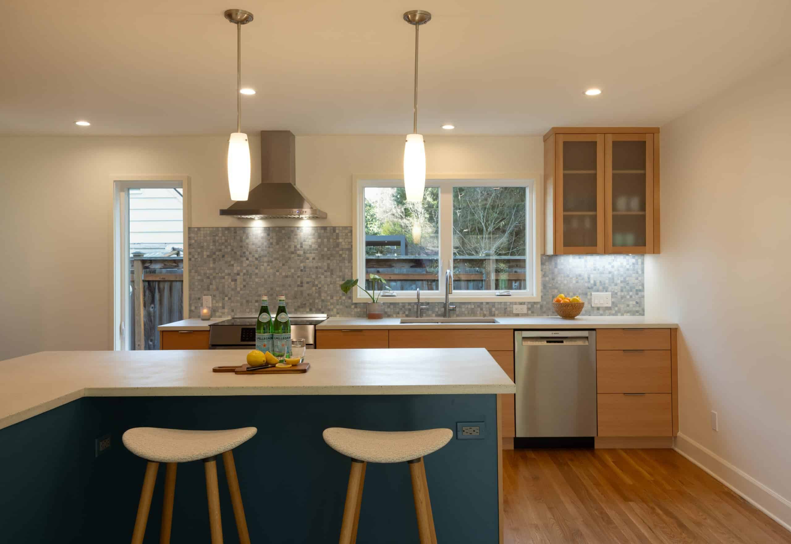 Modern kitchen with wood cabinets, a center island with two stools, stainless steel appliances, pendant lights, and a tile backsplash. Bottled water and lemons are on the island.