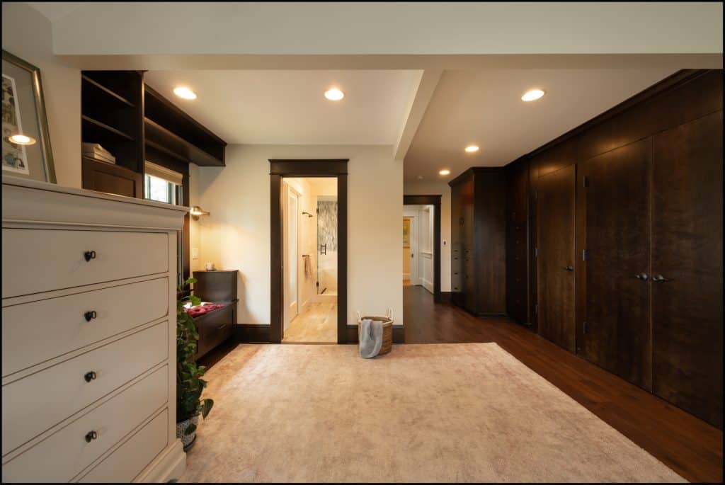 Spacious bedroom with a beige carpet, white dresser, dark wood closets, built-in shelves, and houseplant—all part of an award-winning remodel. An open doorway leads to a bright bathroom for seamless living.