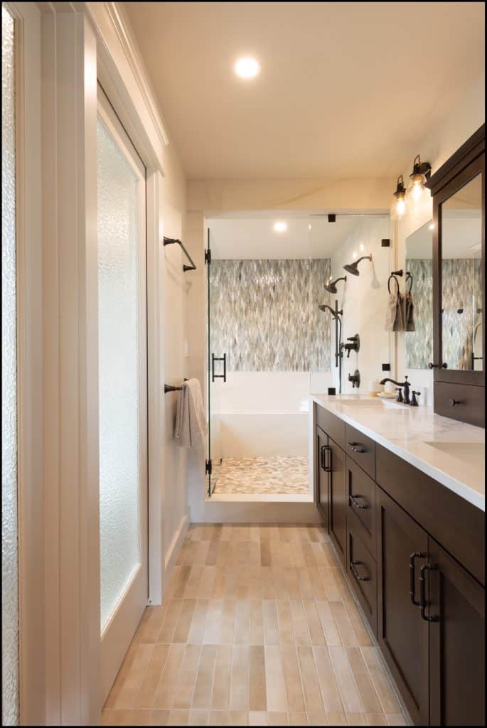Modern bathroom with double dark wood vanity, large mirror, glass shower enclosure, textured tile wall, and neutral floor tiles—an award-winning residential interior by a NARI Remodeler of the Year.