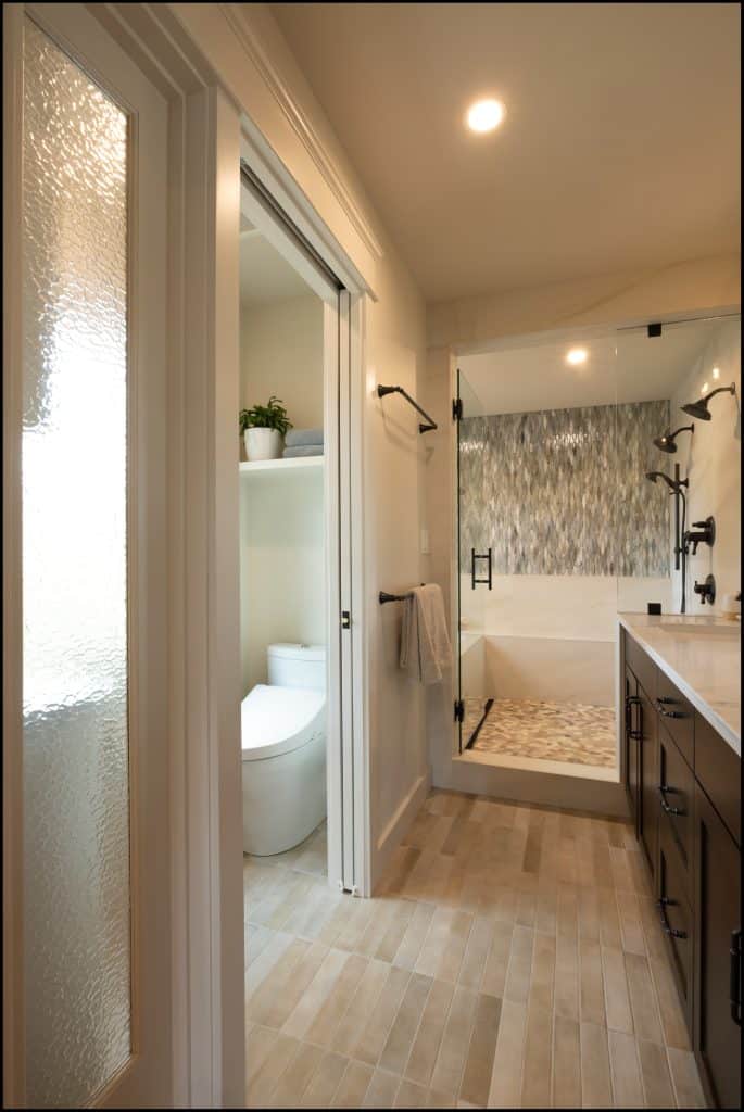 This modern bathroom, part of an award-winning remodel, features a glass-enclosed shower, double sink vanity, towel rack, and a separate toilet area with shelves and a potted plant.