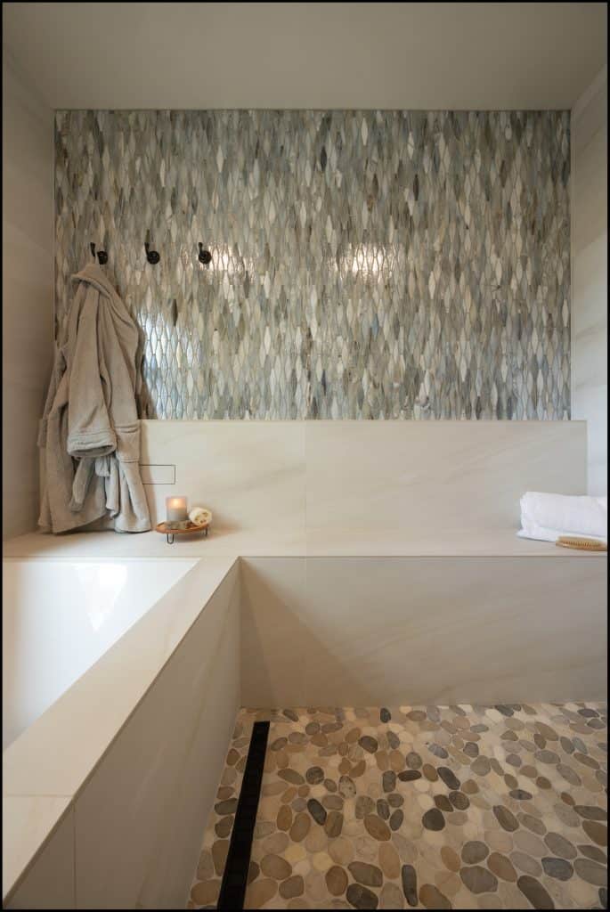 Modern bathroom with a stone tile floor, geometric wall tiles, and a built-in bathtub showcases an award-winning remodel. A folded towel, robes on hooks, and a candle and soap tray by the tub add comfort and style.