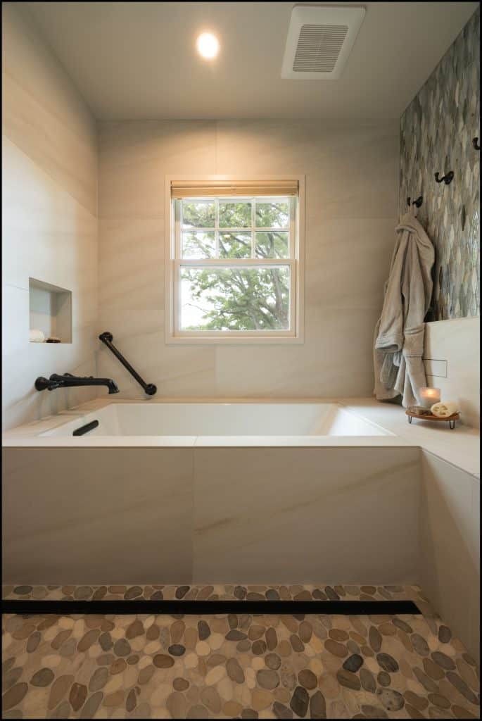 Modern bathroom with a large bathtub, wall-mounted grab bar, window, pebble-style floor, robe on hooks, and a candle on a tray by the tub—an award-winning remodel recognized by NARI.