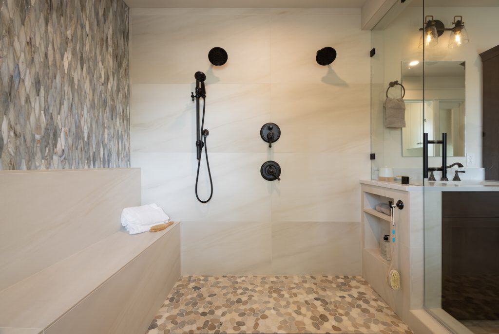 Awarded Remodeler of the Year, this stunning residential interior remodel features a modern walk-in shower with dual black showerheads, a handheld sprayer, built-in bench, pebble tile floor, and glass partition beside a sleek vanity area.